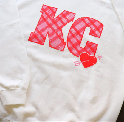 White sweatshirt with red 'KC' lettering and heart design on a gray surface
